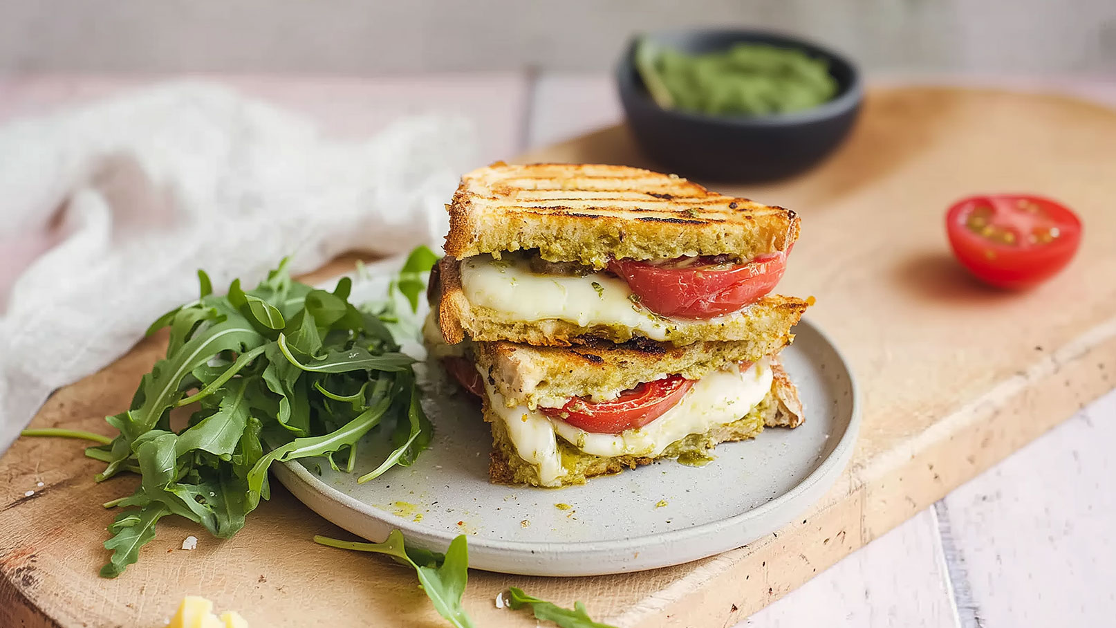 Ein Teller mit einem gegrillten Sandwich, gefüllt mit geschmolzenem Käse und frischen Tomatenscheiben. Daneben liegt ein frischer Salat aus Rucola. Im Hintergrund befindet sich eine kleine Schüssel mit einer grünen Paste.