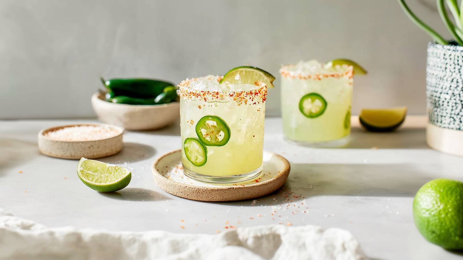 Zwei Gläser Getränk mit Eis, garniert mit grünen Jalapeño-Scheiben und Limetten, stehen auf einem hellen Tisch. Neben den Gläsern liegen Limetten und kleine Schalen mit Salz. Das Getränk wirkt erfrischend und farbenfroh.