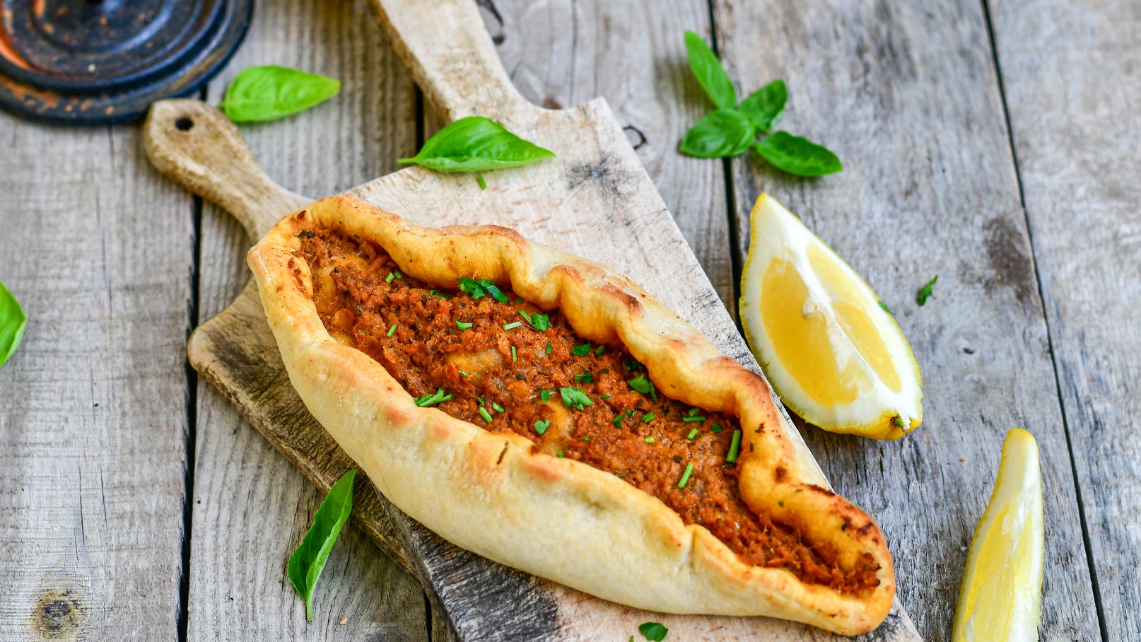 Ein frisch gebackenes Pide, gefüllt mit würziger, brauner Fleischfüllung, liegt auf einem Holzbrett. Neben dem Pide sind frische Zitronenscheiben und einige grüne Basilikumblätter platziert.