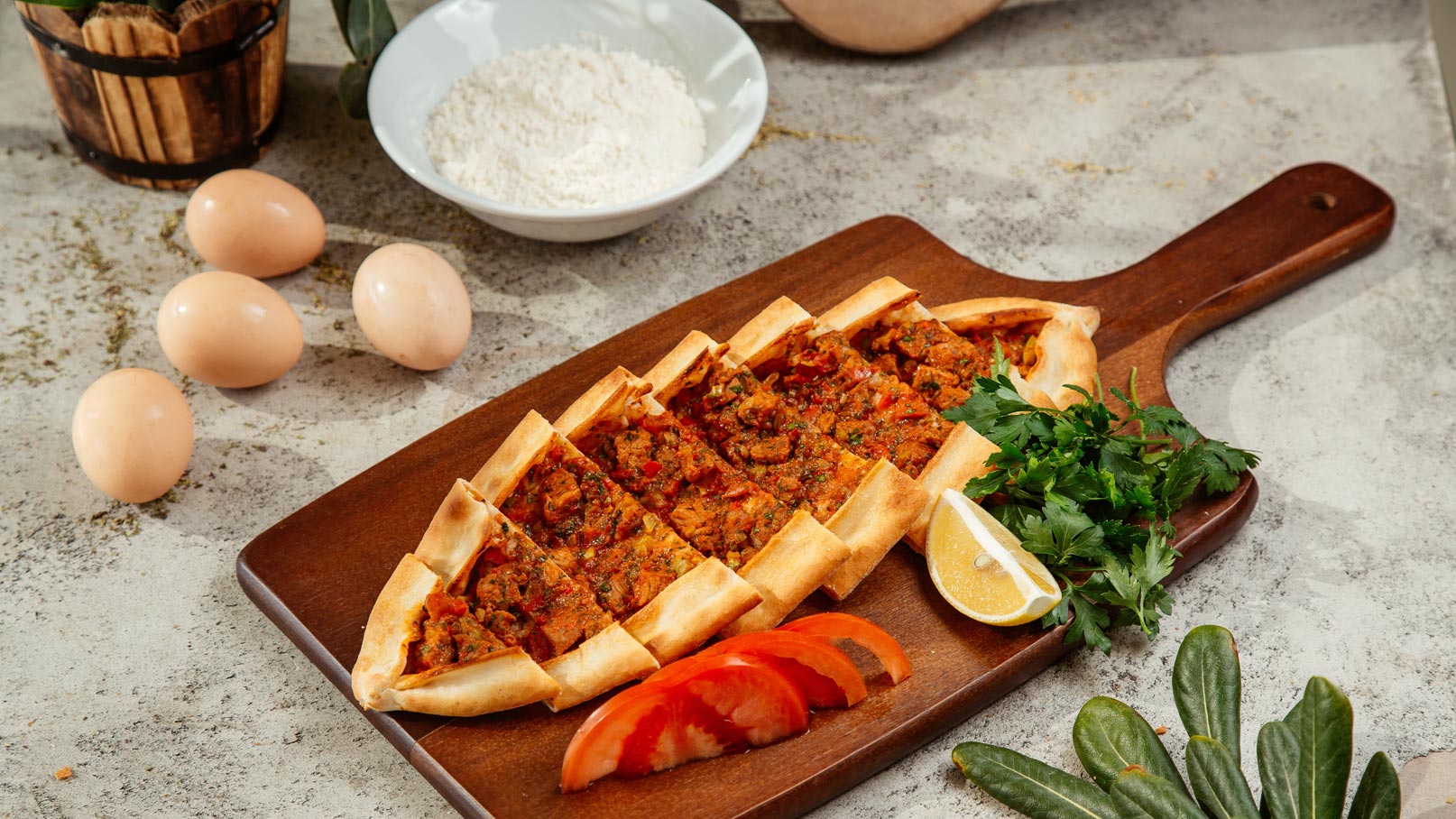 Auf einem Holzbrett liegt eine Portion gefüllte Teigtaschen, geschnitten und garniert mit Tomatenscheiben, Zitronenstück und frischer Petersilie. Im Hintergrund sind einige Eier und eine Schüssel mit Mehl zu sehen.