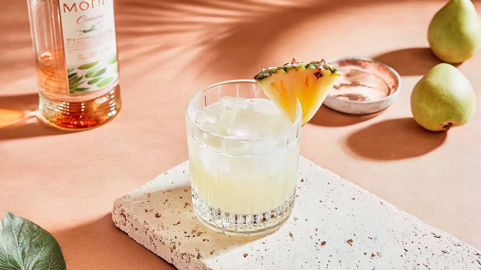 Ein Glas mit einem leichten, erfrischenden Cocktail ist mit Eis gefüllt und mit einer Ananasscheibe dekoriert. Es steht auf einem grauen Untersetzer aus Stein auf einer pastel-braunen Oberfläche. Im Hintergrund sind eine Flasche Likör und einige grüne Birnen sichtbar.