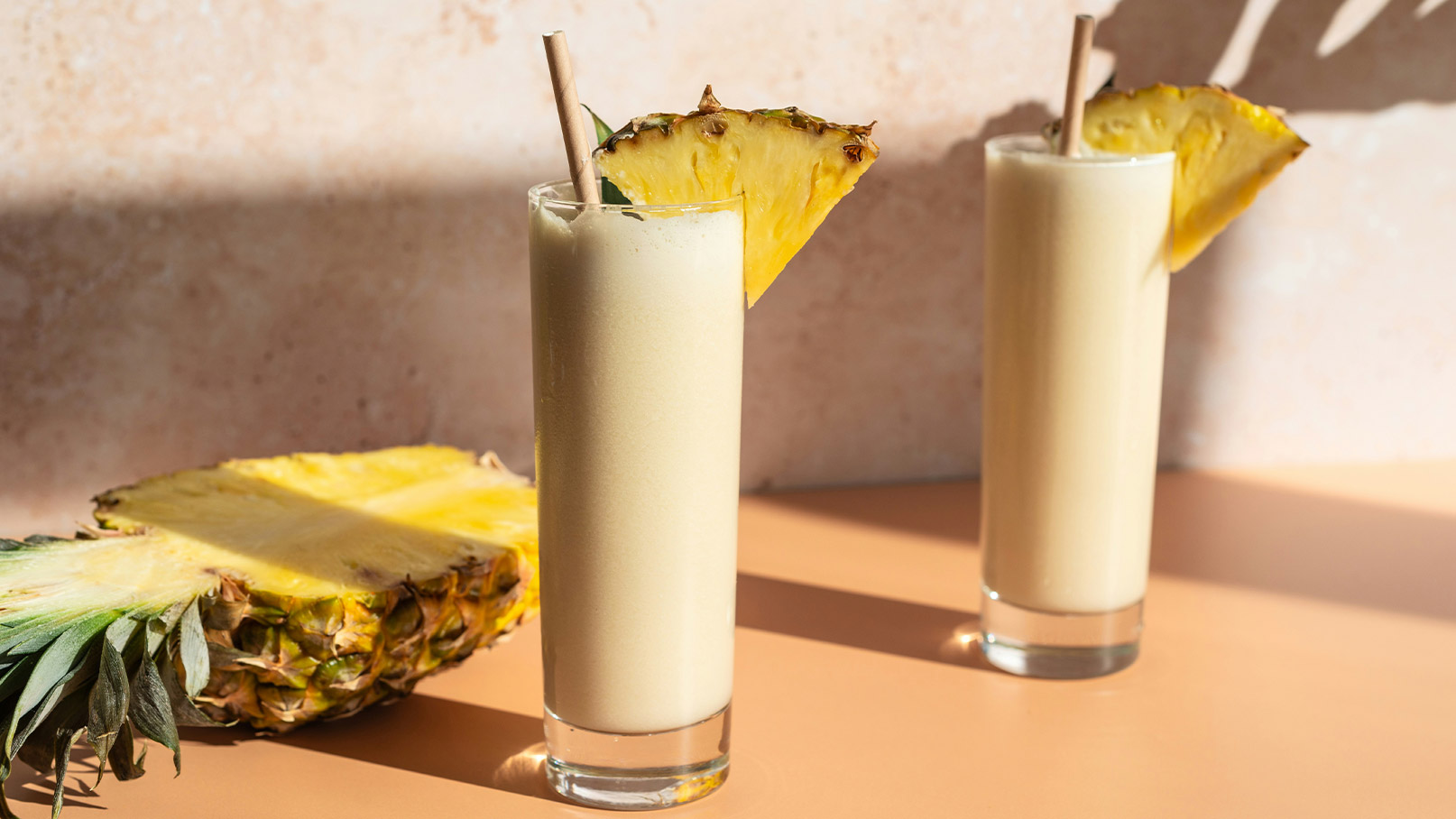 Zwei hohe Gläser mit einem cremigen Ananasgetränk, verziert mit einem Ananasstück und einem Strohhalm. Im Hintergrund liegt eine halbierte Ananas auf einem hellen Tisch. Die angenehme, sonnige Atmosphäre lässt das Getränk frisch und einladend erscheinen.