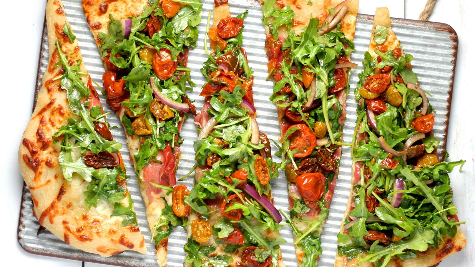 Eine große flache Pizza mit knusprigem Rand, belegt mit frischem Rucola, getrockneten Tomaten und roten Zwiebeln. Die Pizza ist in gleichmäßige Stücke geschnitten und auf einem gestreiften Blech angerichtet, was einen appetitlichen Anblick bietet.
