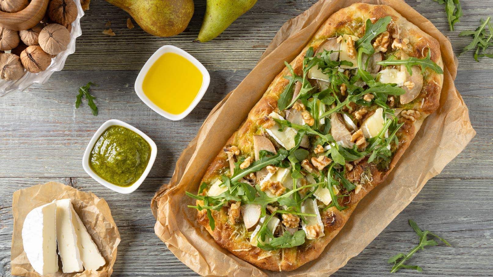 Ein frisch gebackenes Fladenbrot, belegt mit blättrigem Käse, Birnenscheiben, Rucola und Walnüssen. Neben dem Fladenbrot befinden sich kleine Schalen mit grünem Pesto und Olivenöl. Im Hintergrund liegen einige Walnüsse und Birnen. Der Untergrund ist aus Holz.