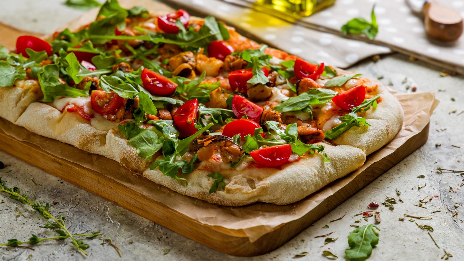 Ein frisch gebackenes Pizza-Brot liegt auf einem Holzbrett. Belegt mit geschmolzenem Käse, Tomatenscheiben und frischem Rucola, zeigt es eine bunte Mischung aus Zutaten. Im Hintergrund sind ätherische Kräuter und eine Flasche Olivenöl sichtbar, die die Präsentation der Pizza ergänzen.
