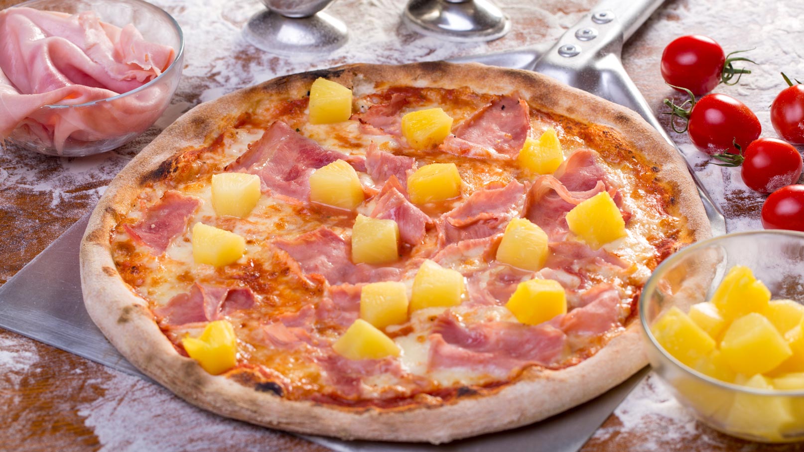Eine frisch gebackene Pizza mit Schinken und Ananas, garniert mit Basilikum. Die Pizza hat einen knusprigen Rand und geschmolzenen Käse. Im Hintergrund liegen frisches Gemüse wie Ananas und Tomaten.