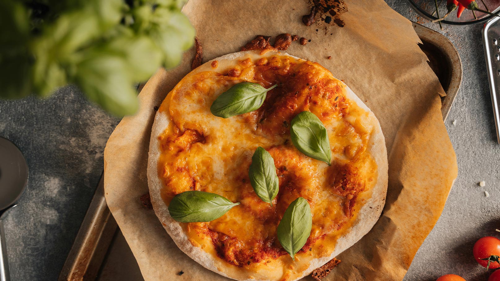 Ein frisch gebackener Pizza mit geschmolzenem Käse, garniert mit frischem Basilikum, liegt auf Backpapier. Die Pizza hat eine goldbraune Kruste und ist auf einem rustikalen Tisch arrangiert. Kleine Tomaten und Küchenutensilien sind im Hintergrund sichtbar.