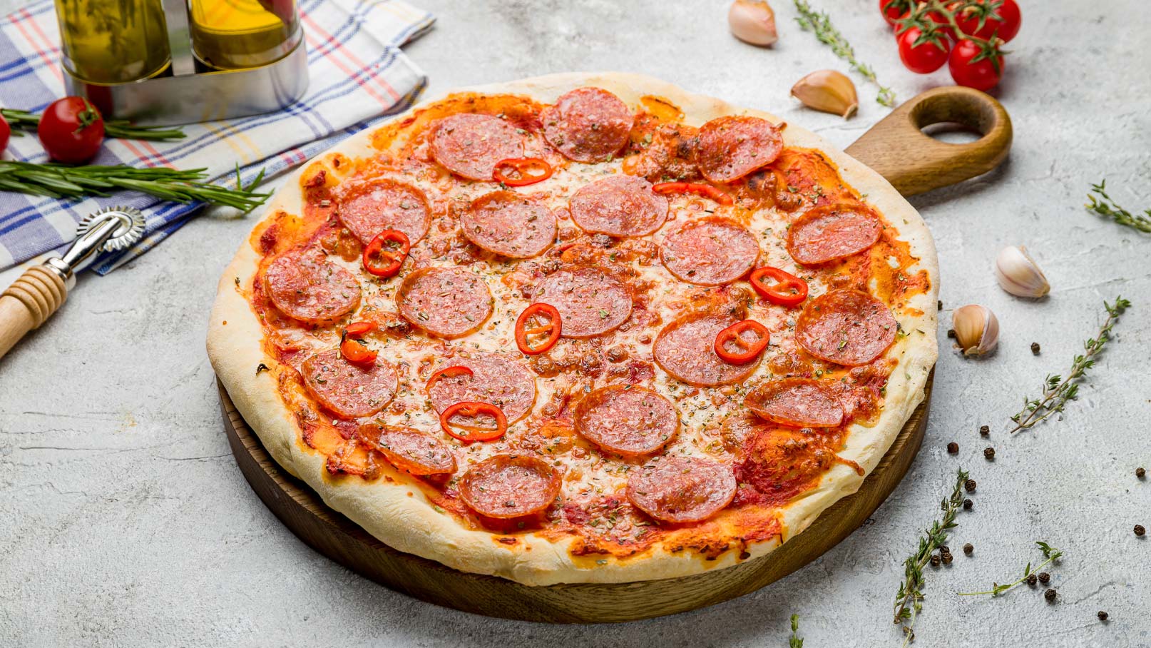 Eine frisch gebackene Pizza liegt auf einem runden Holzbrett. Sie ist mit Tomatensoße, geschmolzenem Käse, zahlreichen Peperoni-Scheiben und roten Chilischoten belegt. Rund um die Pizza sind einige Kräuter, Knoblauchzehen und kleine Tomaten platziert.