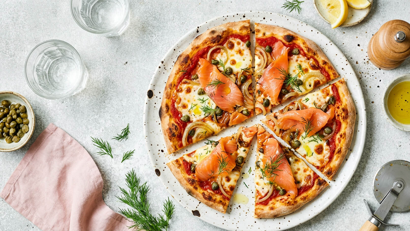 Eine frisch gebackene Pizza mit Tomatensauce, Käse, Zwiebeln, Räucherlachs und Kapern, großzügig mit frischem Dill garniert. Die Pizza ist in Stücke geschnitten und auf einem weißen Teller angerichtet, umgeben von kleinen Schalen mit Kapern und Olivenöl.