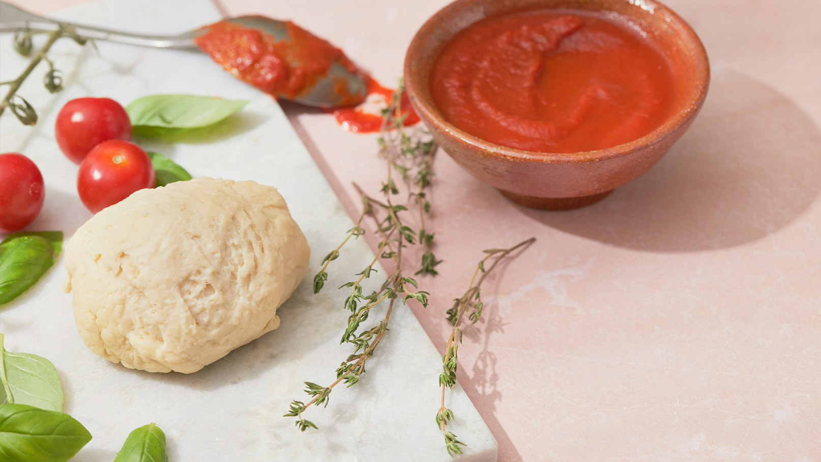 Auf einer Arbeitsfläche liegt ein Klumpen Teig, umgeben von frischen Basilikumblättern und Kirschtomaten. Neben dem Teig steht eine Schüssel mit Tomatensoße, aus der ein Löffel herausragt. Thymianzweige sind ebenfalls verstreut.