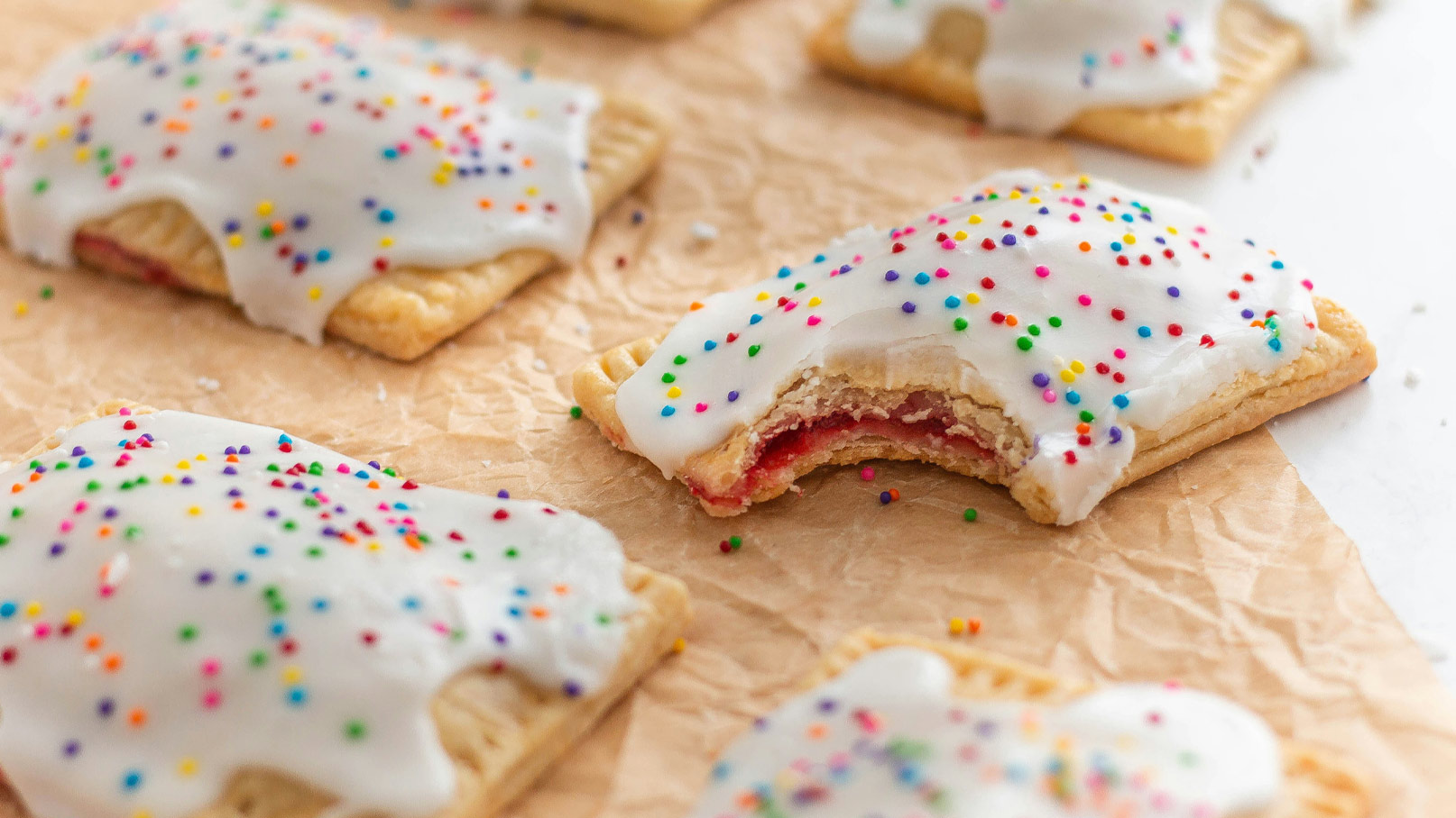 Eine Auswahl von fruchtigen Plätzchen mit einer weißen Zuckerglasur und bunten Streuseln. Eines der Plätzchen ist halb aufgegessen, sodass die fruchtige Füllung sichtbar ist. Die Plätzchen liegen auf braunem Papier.