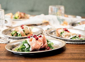 Auf einem Holztisch stehen mehrere silberne Teller mit frischen Salatgerichten. Jedes Gericht enthält Rucola, umwickelt mit Schinken und garniert mit Granatapfelkernen. Der Hintergrund zeigt verschwommene Gläser und weitere Teller.