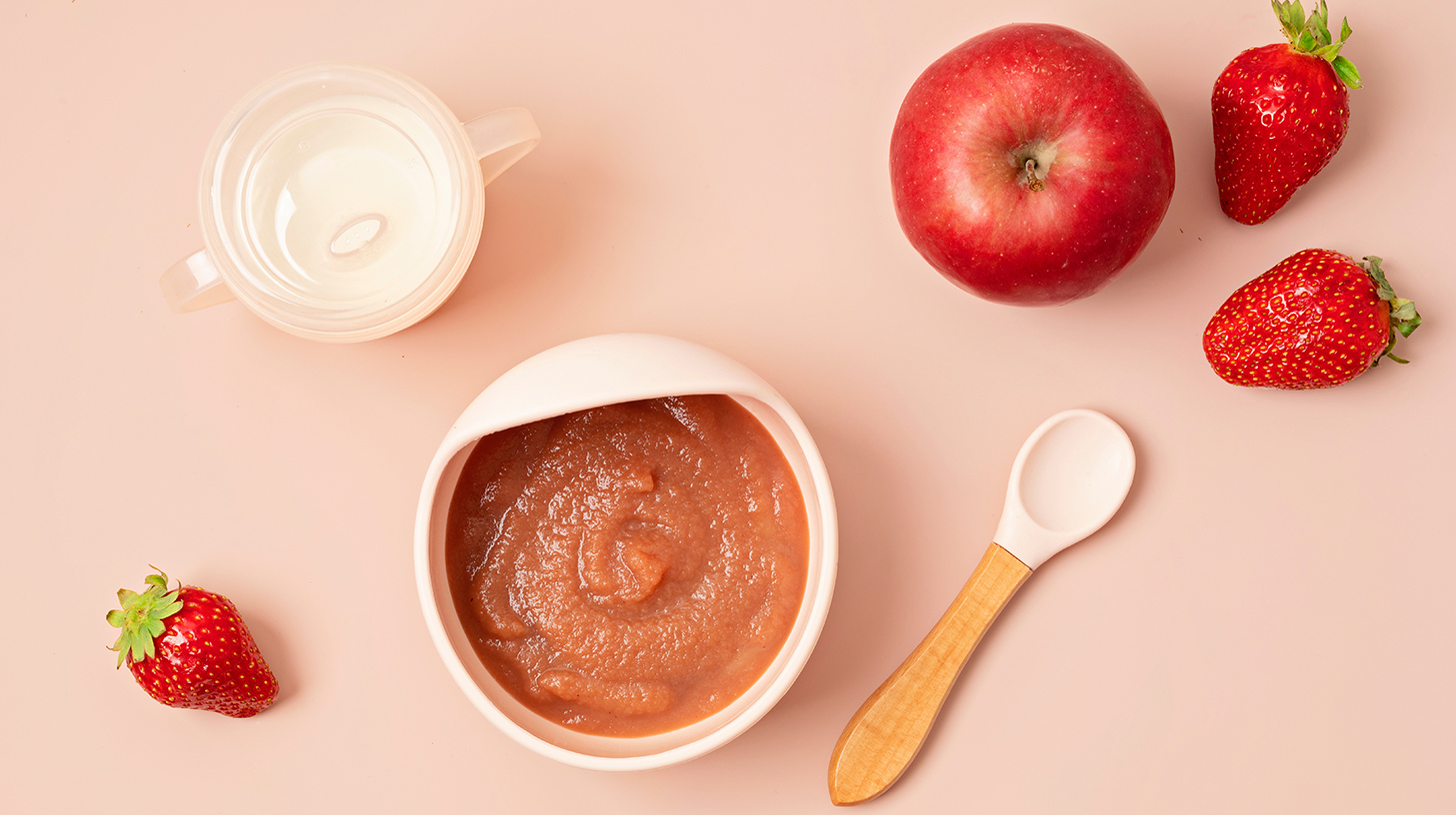 Eine Schüssel mit Apfelmus steht auf einer rosa Fläche. Daneben liegen ein roter Apfel, mehrere Erdbeeren und ein Löffel aus Holz. Ein kleiner Becher mit Wasser befindet sich ebenfalls in der Nähe.