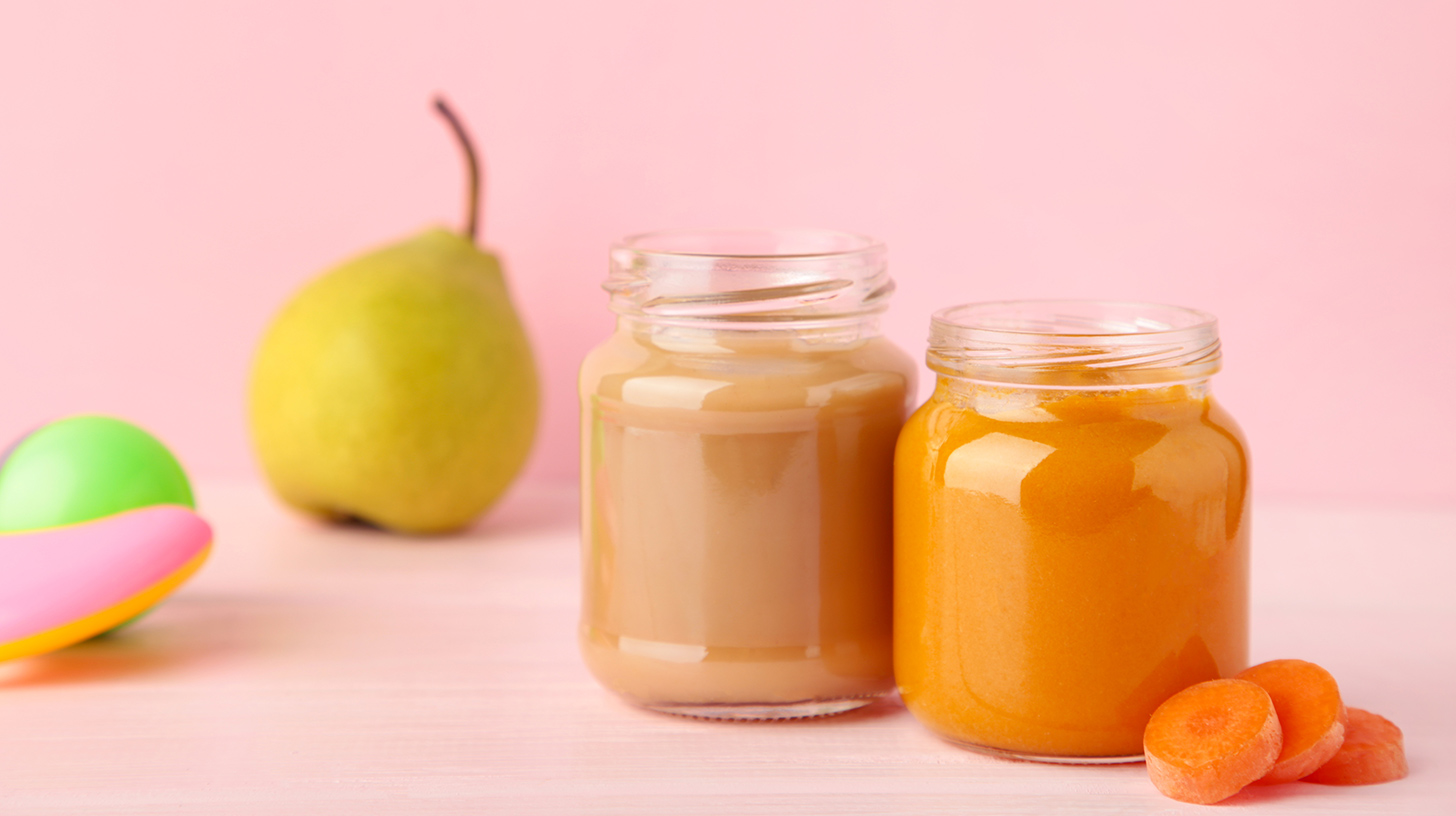 Zwei Gläser mit Babynahrung stehen auf einem hellen Tisch. Das linke Glas enthält eine bräunliche Mischung, das rechte Glas eine orangefarbene Mischung. Neben dem rechten Glas liegen einige orangefarbene Karottenstücke. Im Hintergrund ist eine grüne Birne sichtbar, vor einem rosa Hintergrund.