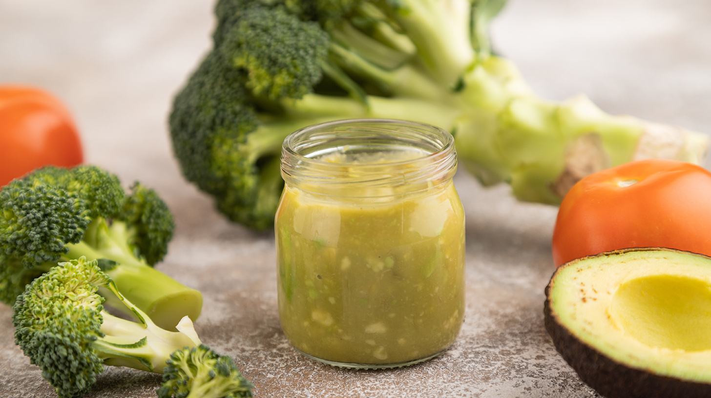 Ein Glas mit grüner Sauce oder Püree steht im Vordergrund, umgeben von frischem Brokkoli, einer geschnittenen Avocado und einer Tomate. Die Zusammensetzung deutet auf gesunde, pflanzliche Zutaten hin.