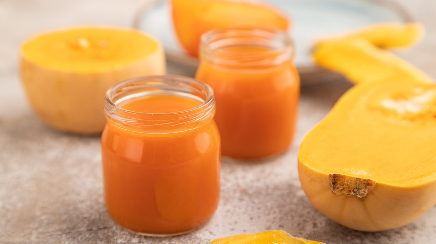 Zwei kleine Gläser mit orangefarbenem Kürbissaft stehen auf einer Tischoberfläche. Im Hintergrund liegt ein halbierter Hokkaido-Kürbis. Der Saft hat eine lebendige Farbe und der Kürbis hat eine glatte, goldene Oberfläche.