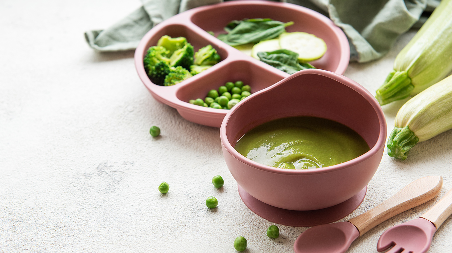 Ein rosa Schüsselchen mit grüner Gemüsesuppe steht auf einem Tisch. Daneben liegt ein Teller mit verschiedenen Gemüsesorten wie Brokkoli, Spinat und Zucchini. Zwei Holzlöffel und ein paar grüne Erbsen sind ebenfalls sichtbar.
