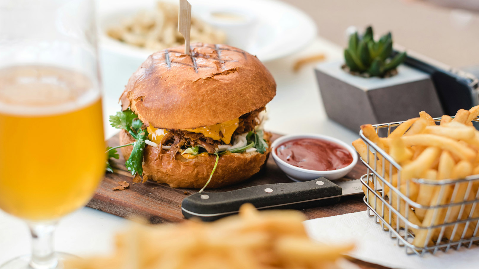 Ein saftiger Burger mit gegrilltem Brötchen, gefüllt mit zerfällt Fleisch, Käse und frischen Zutaten, steht auf einem Holzbrett. Daneben ein kleiner Behälter mit Ketchup und eine Portion Pommes in einem Korb. Im Hintergrund ein Glas Bier.