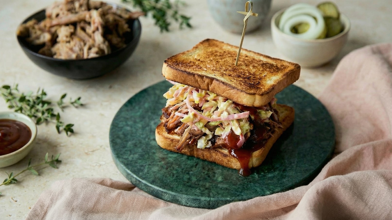 Ein Sandwich aus geröstetem Brot, gefüllt mit zartem Pulled Pork, Krautsalat und BBQ-Sauce. Es ist auf einem grünen Teller angerichtet. Im Hintergrund ist eine Schüssel mit weiteren Fleischstücken zu sehen sowie einige Garnituren, die das Gericht begleiten.