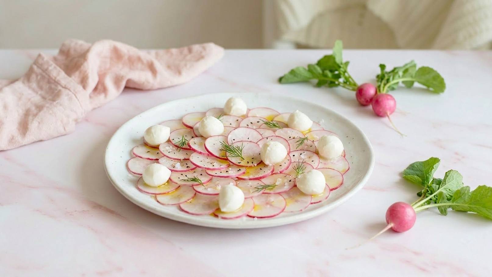 Ein Teller mit dünn geschnittenen Radieschen, kunstvoll in einer spiralförmigen Anordnung. Frische Kräuter sind darüber gestreut. Neben dem Teller liegen einige ganze Radieschen und ein rosa Tuch. Der Hintergrund ist hell und harmonisch.