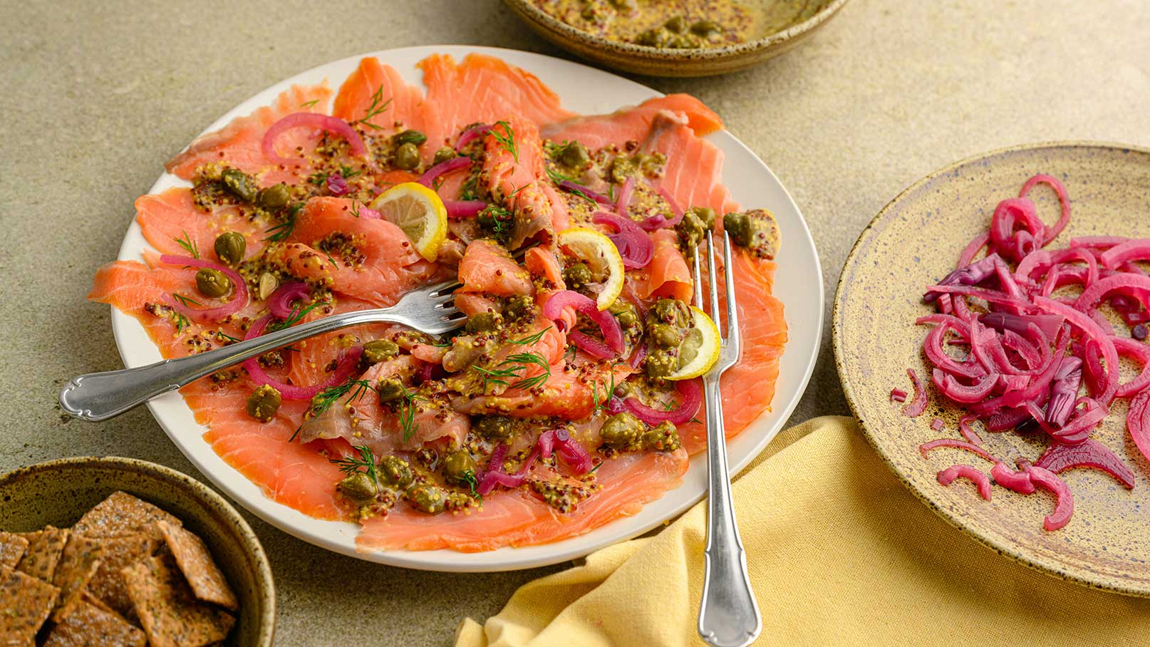 Ein Teller mit dünn geschnittenem Lachs, belegt mit Kapern, Senf und roten Zwiebeln. Neben dem Teller liegen zwei Gabeln. Daneben ist ein weiterer Teller mit geschnittenen roten Zwiebeln und eine Schale mit knusprigem Brot.