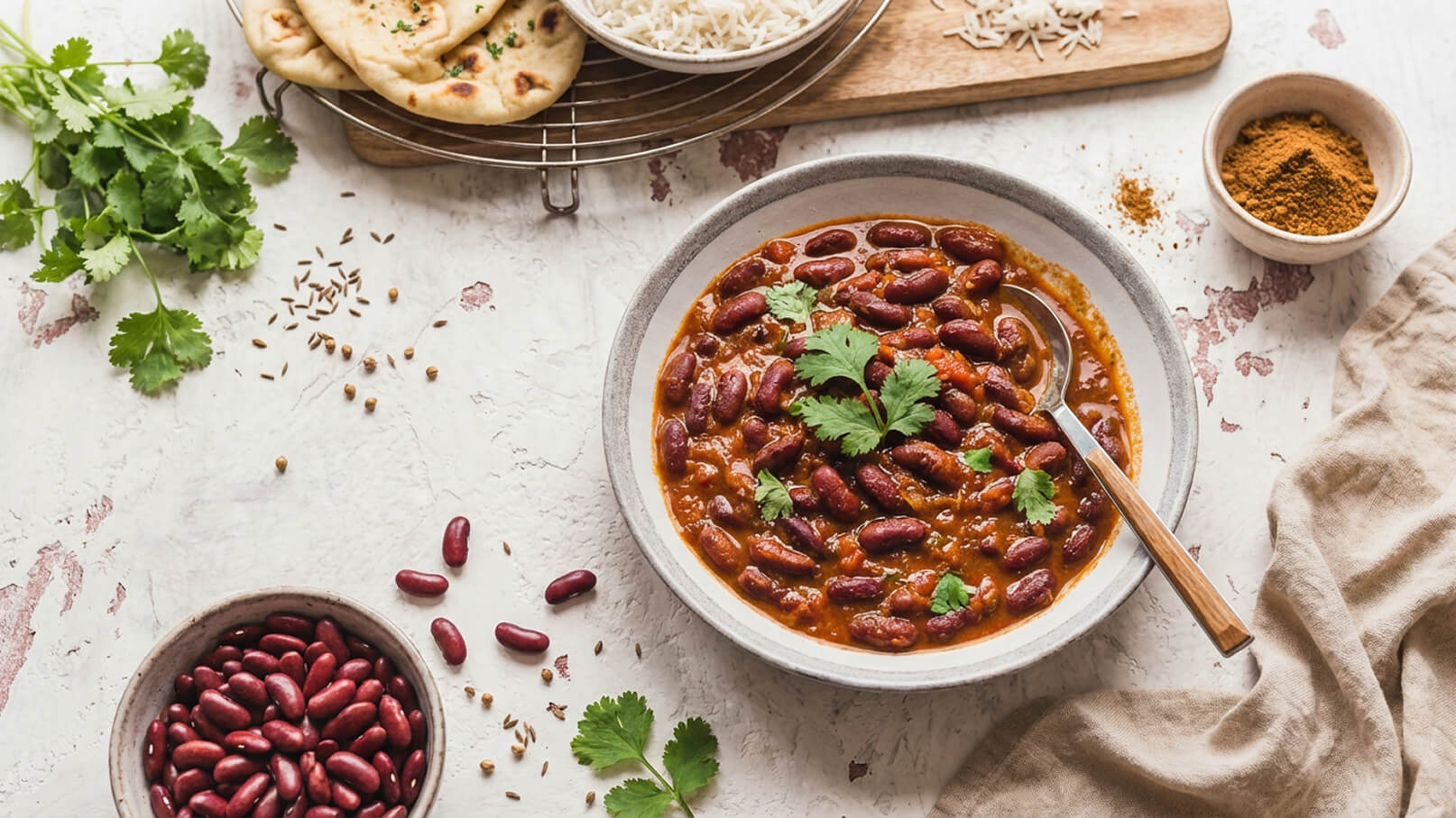 Eine Schüssel mit rotem Bohnen-Curry, garniert mit frischem Koriander, steht im Vordergrund. Daneben liegt eine Schale mit roten Bohnen. Im Hintergrund sind frisch gebackenes Naan-Brot und eine Portion Reis. Gewürze sind ebenfalls sichtbar.
