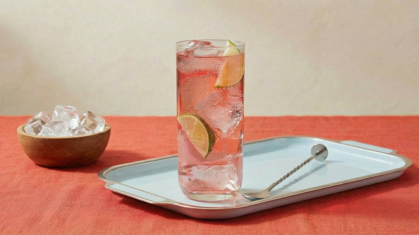 Ein hohes Glas mit einem sprudelnden Getränk, gefüllt mit Eiswürfeln und Limetten- sowie Zitronenscheiben, steht auf einem blauen Tablett. Daneben befindet sich eine Schüssel mit weiteren Eiswürfeln auf einem roten Tisch.
