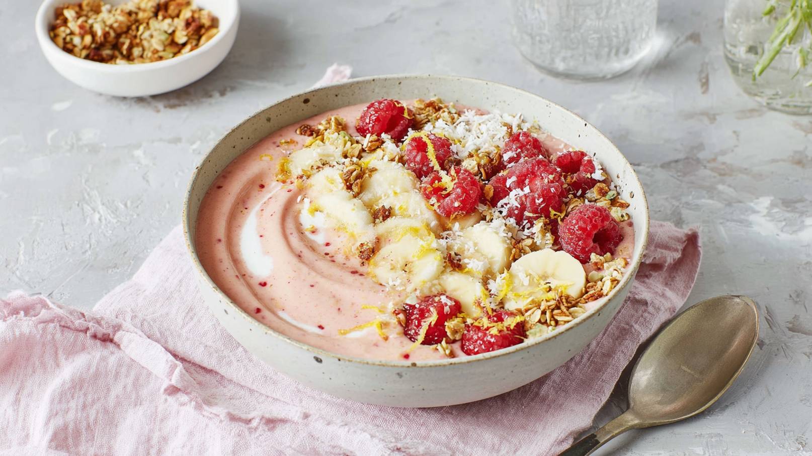 Eine Schüssel mit fruchtigem Joghurt, garniert mit Himbeeren, Bananenscheiben, Kokosraspeln und Müsli. Der Joghurt hat eine rosa Farbe und ist mit einem Schuss Sahne verziert. Die Schüssel steht auf einem rosa Tuch, daneben liegt ein Löffel.