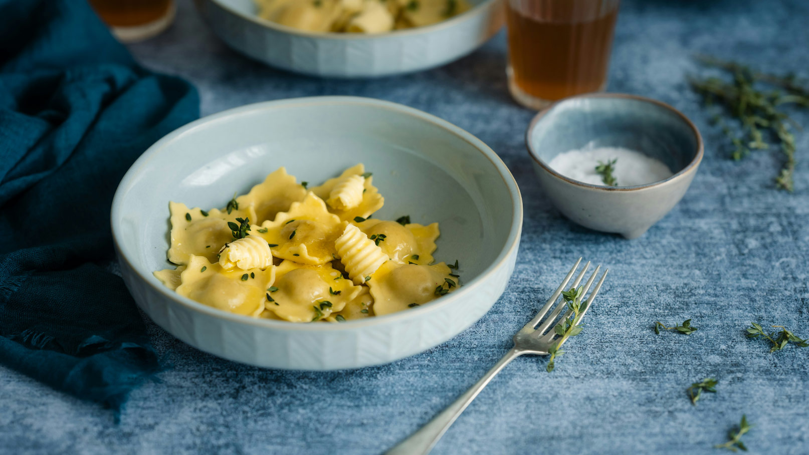 Eine blaue Schüssel mit frischen Ravioli, die mit Butter und frischen Kräutern garniert sind. Daneben steht eine kleine Schüssel mit einer cremigen Sauce. Im Hintergrund sind weitere Ravioli und ein Getränk zu sehen.
