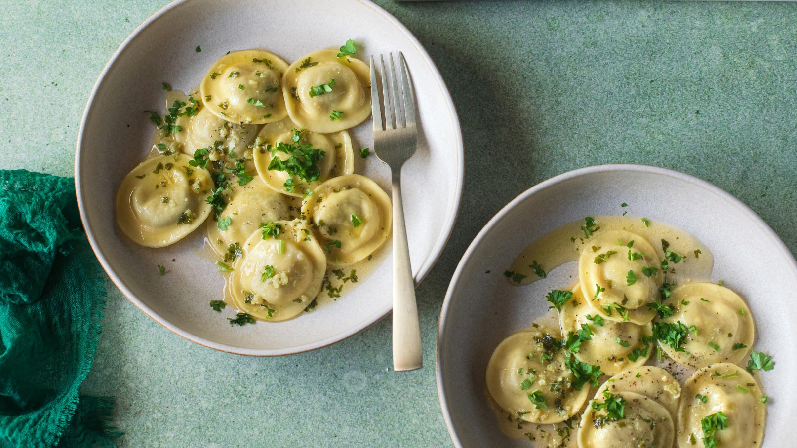 Zwei Teller Ravioli in einer hellen Sauce, garniert mit frischen Kräutern. Ein Teller hat eine Gabel daneben. Die Ravioli sind rund und leicht gefüllt. Der Hintergrund ist in sanften Grüntönen gehalten.