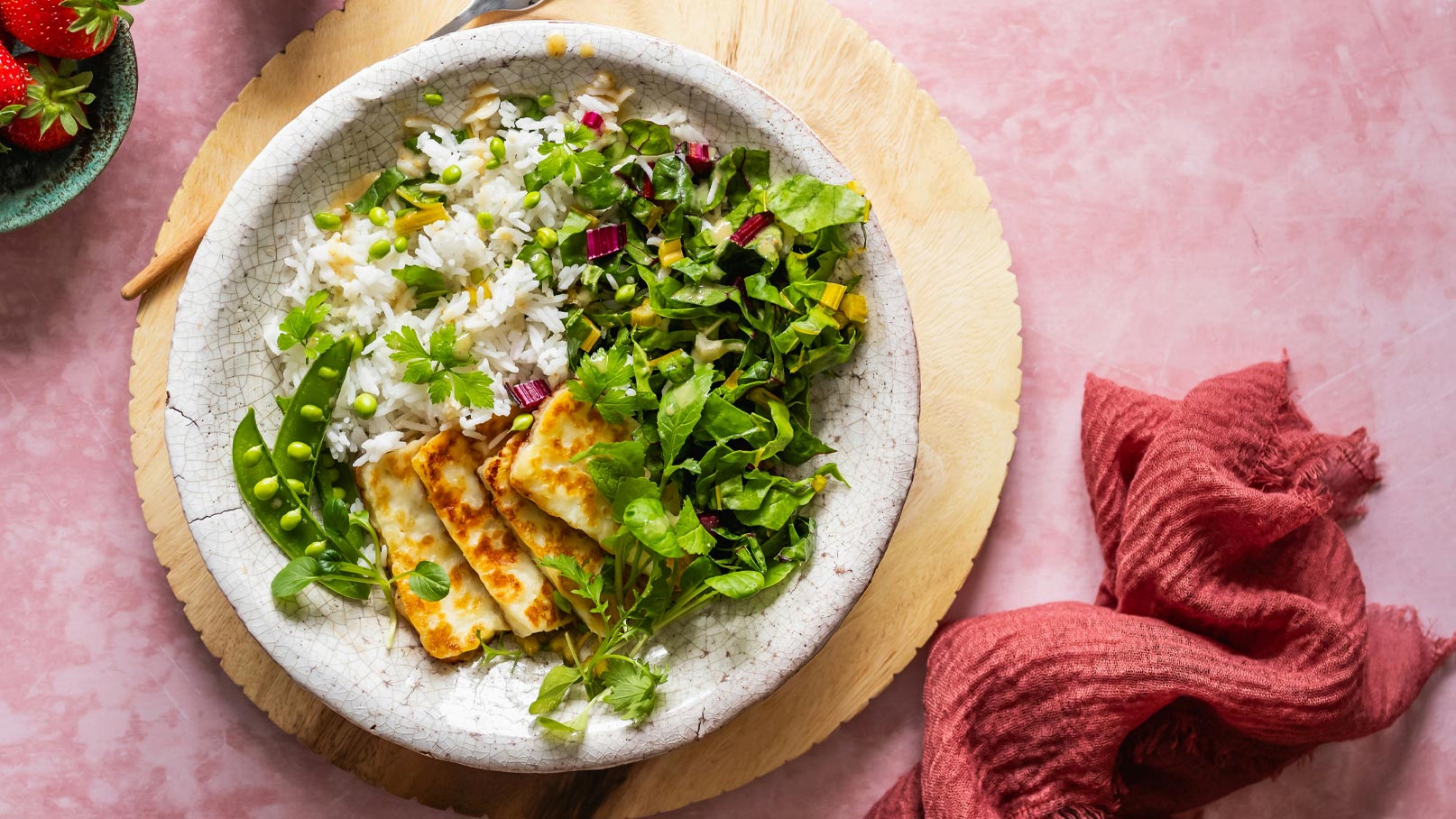 Eine Schüssel mit Reis, grünen Erbsen und frischem, gehacktem Salat. Gegrillte Käsescheiben sind daneben angerichtet. Das Gericht ist auf einem Holzbrett platziert, umgeben von einer rötlichen Serviette und einigen Erdbeeren im Hintergrund.