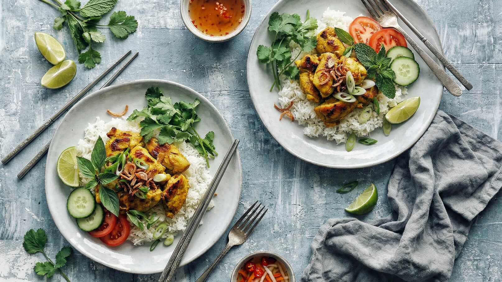 Zwei Teller mit Reis, gegrilltem Hühnchen und frischem Gemüse wie Gurken und Tomaten. Die Gerichte sind mit Koriander und Limettenscheiben garniert. Dazu gibt es eine kleine Schale mit einer würzigen Sauce. Die Teller sind auf einem blauen Tisch angerichtet.