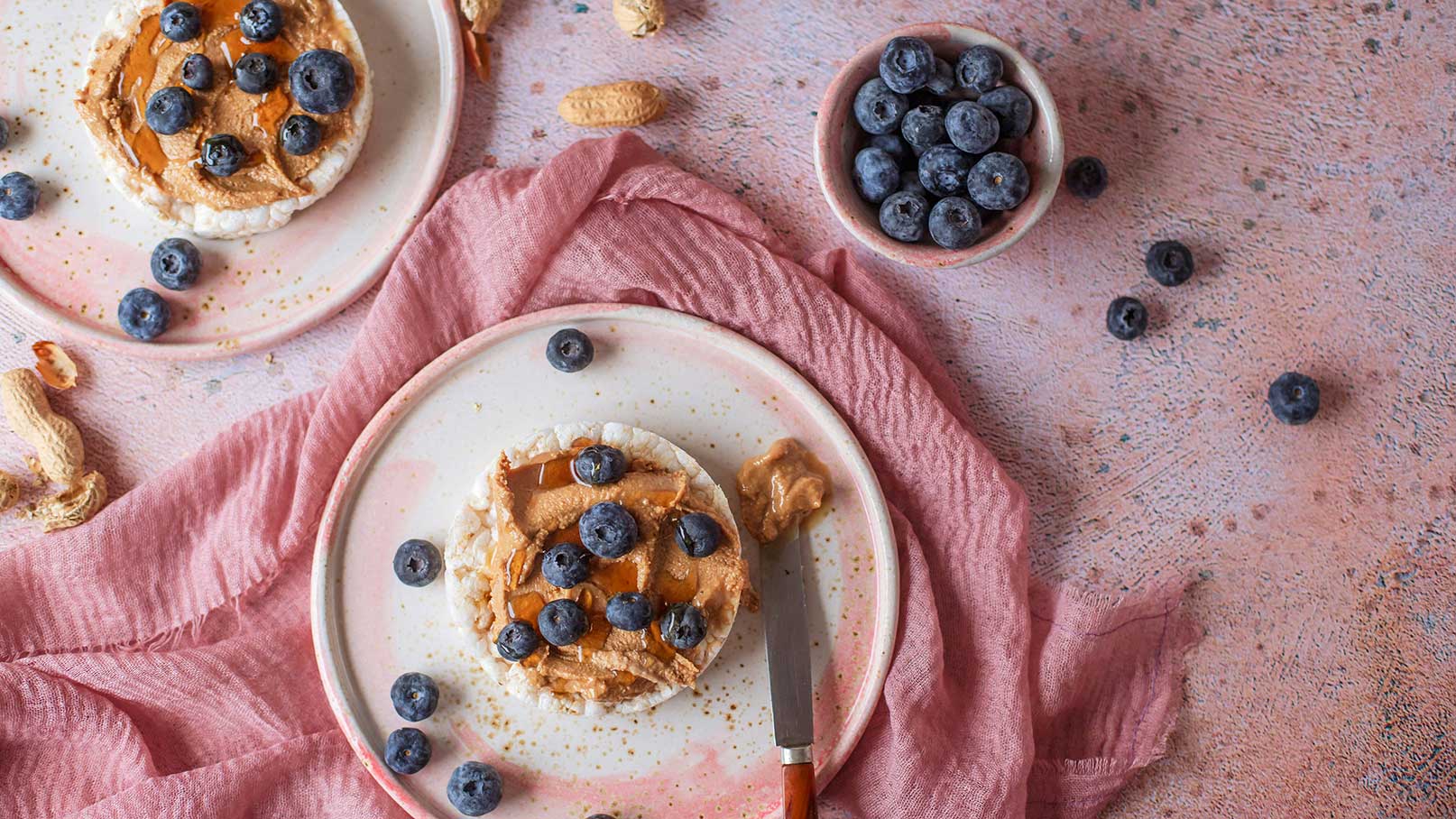 Zwei Teller mit Reiswaffeln, die mit Erdnussbutter und Heidelbeeren belegt sind. Eine Waffel ist teilweise mit einer Gabel auf einem Teller angerichtet. Neben den Tellern liegen einige lose Heidelbeeren. Ein pinkfarbenes Tuch ist sichtbar.