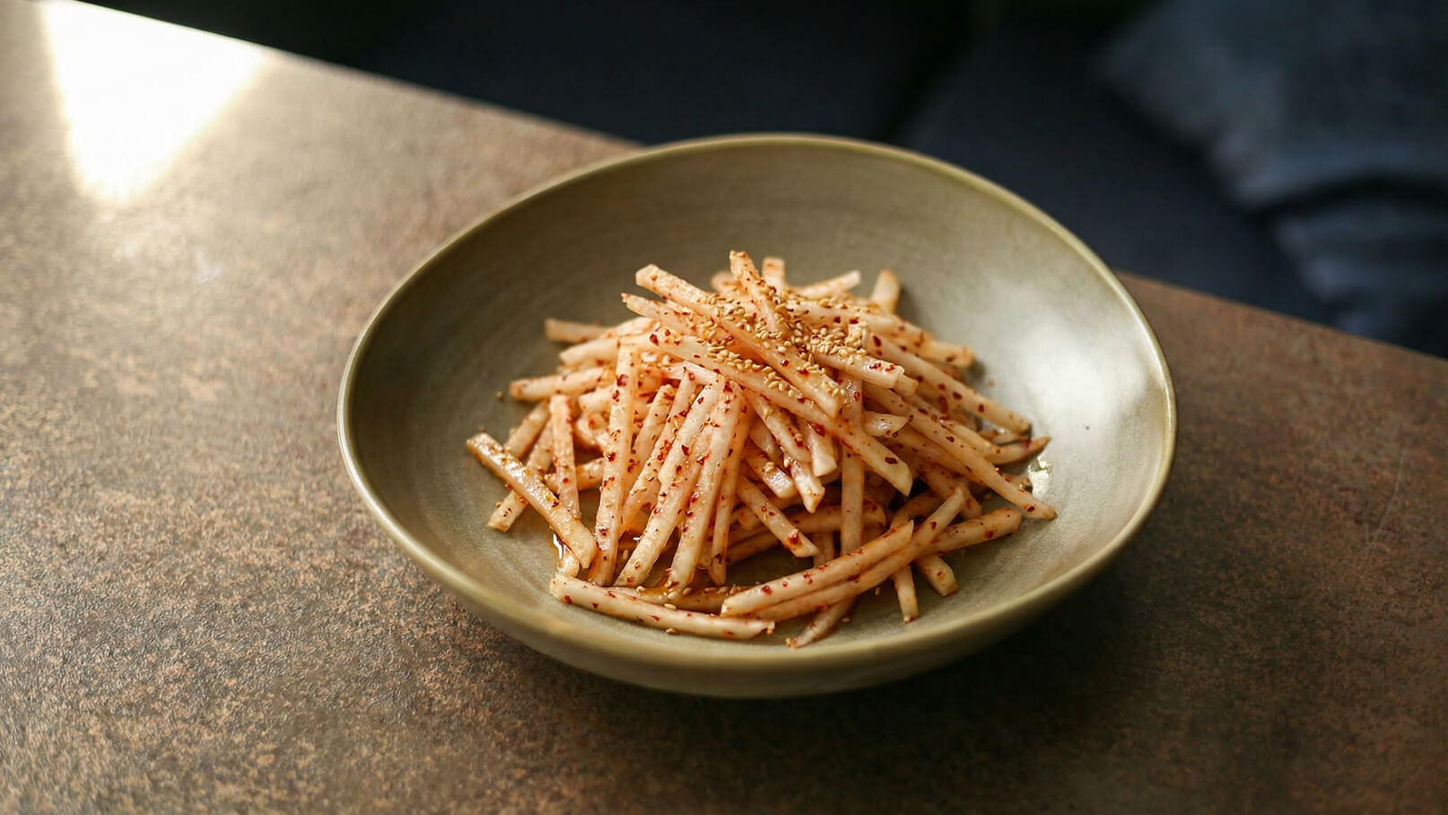 Eine Schüssel mit einer Portion Julienne-Gemüse, fein geschnitten und leicht gewürzt. Die Stücke sind gleichmäßig verteilt und mit einer Prise Sesamsamen bestreut. Die Farbe der Zutaten ist hell und frisch. Der Teller hat eine sanfte, grüne Oberfläche.