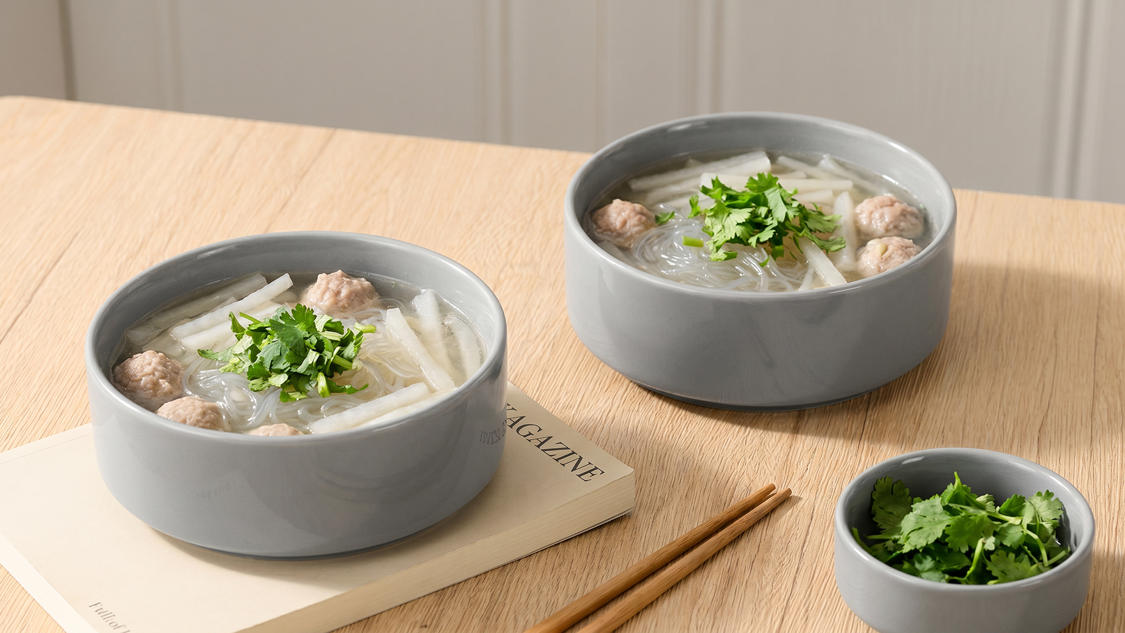 Zwei graue Schalen mit Suppe stehen auf einem Holzstamm. Die Suppe enthält Glasnudeln, Fleischbällchen und frische Kräuter. Neben den Schalen liegt ein kleiner Schälchen mit Kräutern und Essstäbchen.