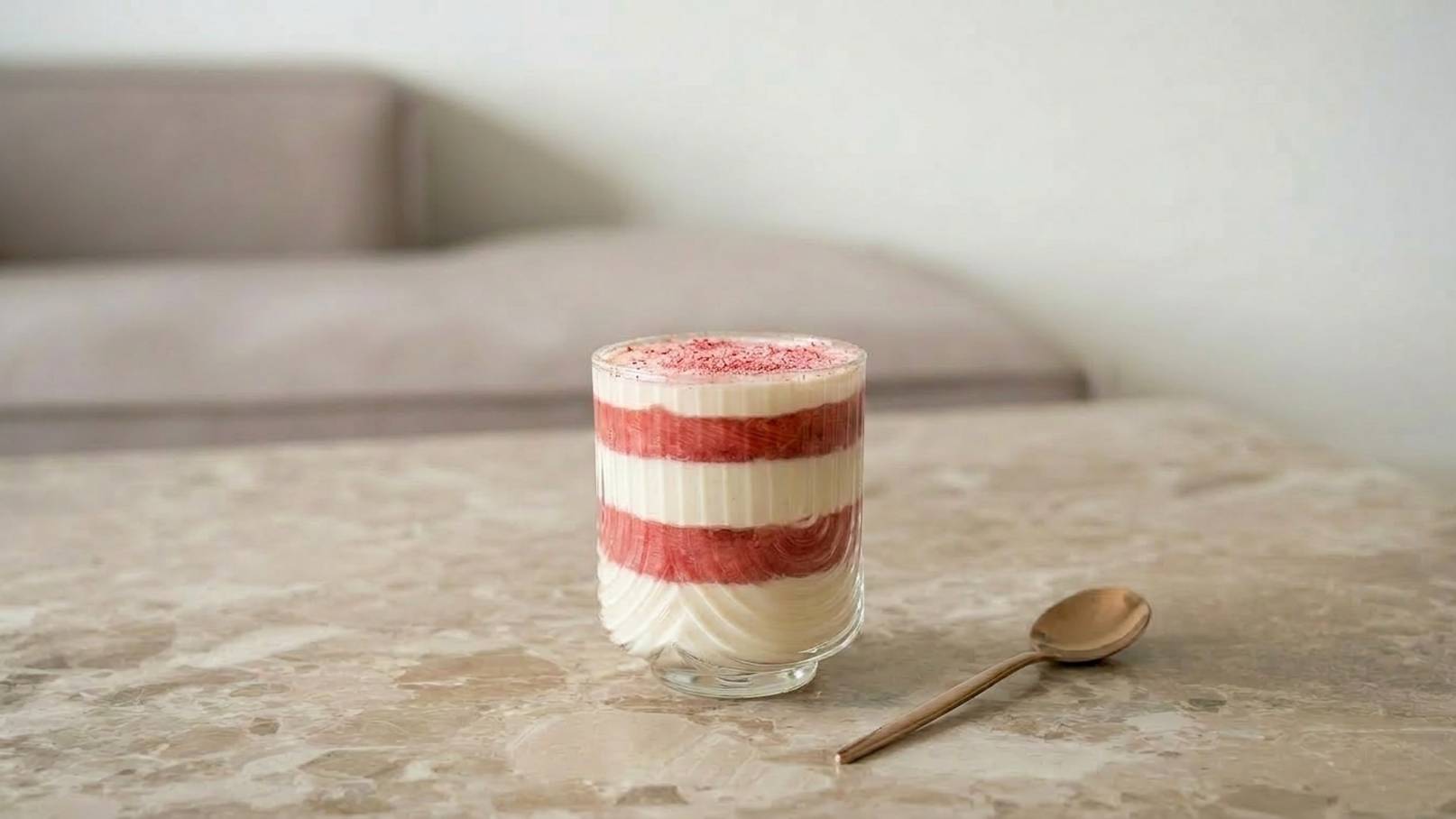 Ein Glas mit Schichtdessert, bestehend aus weißen und roten Schichten, steht auf einem Tisch. Daneben liegt ein kleiner Löffel. Im Hintergrund ist eine bequeme Couch sichtbar.