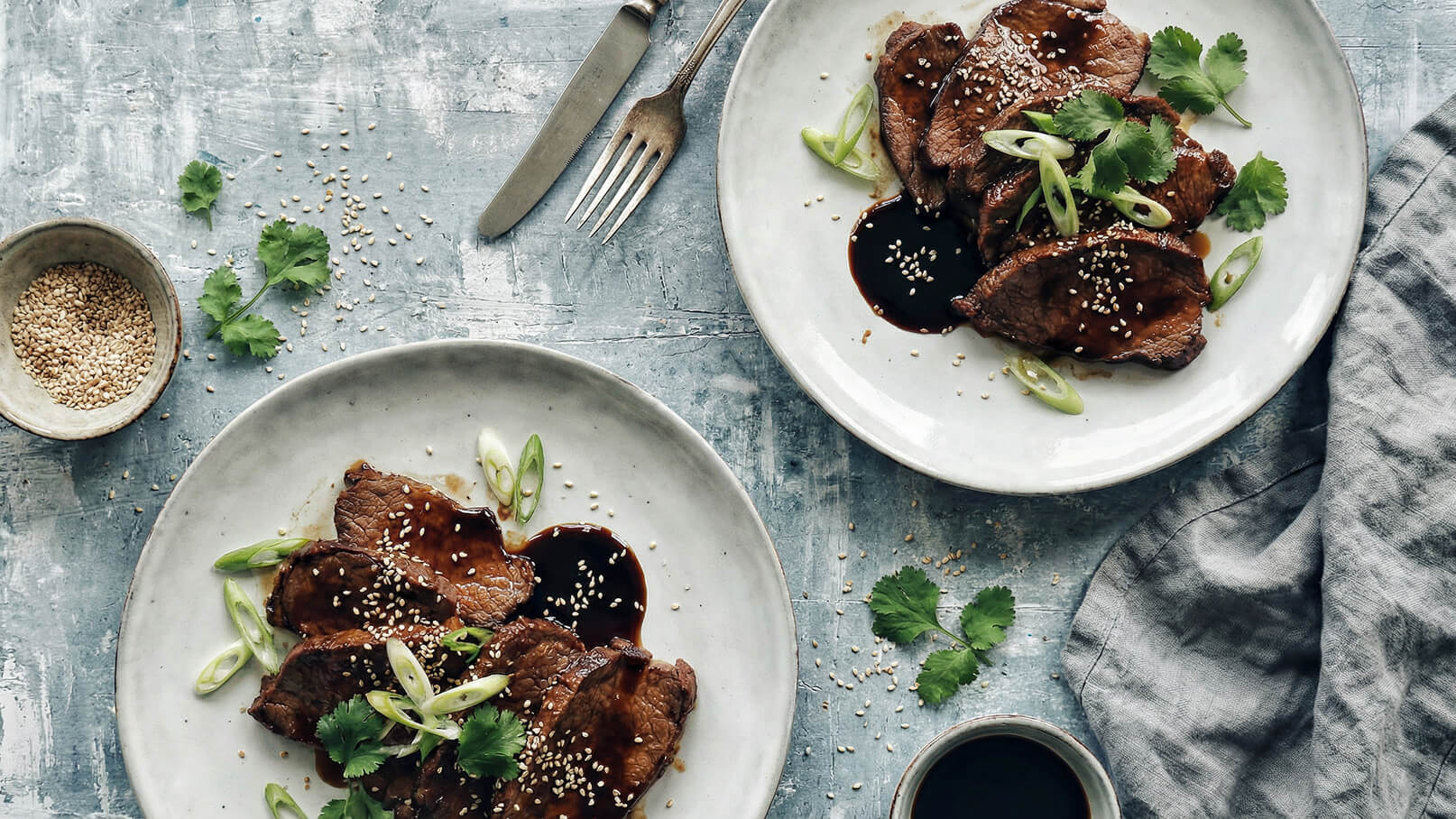 Zwei Teller mit zartem Fleisch in einer dunklen Sauce, garniert mit Frühlingszwiebeln, Sesam und frischem Koriander. Daneben eine kleine Schüssel mit Sesamsamen und ein Glas mit einer dunklen Flüssigkeit. Der Hintergrund ist in einem sanften Blau gehalten.