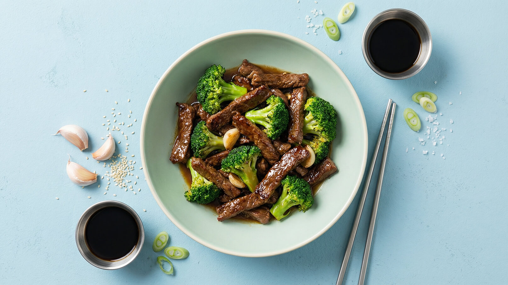 Ein grüner Teller mit Rindfleischstreifen und Brokkoli in einer dunklen Soße. Knoblauchzehen und Sesamsamen liegen daneben. Einige geschnittene Frühlingszwiebeln und eine kleine Schale mit dunkler Soße sind ebenfalls sichtbar. Die Hintergrundfarbe ist hellblau.