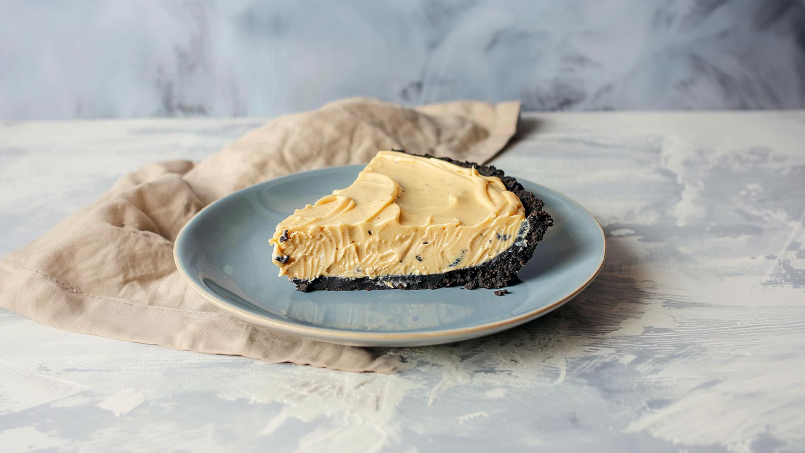 Ein Stück cremiger Kaffeekuchen mit einer schwarzen Keksbodenbasis liegt auf einem blauen Teller. Der Kuchen ist glatt und hat eine helle, beige Farbgebung. Im Hintergrund ist ein weicher, neutraler Stoff sichtbar.