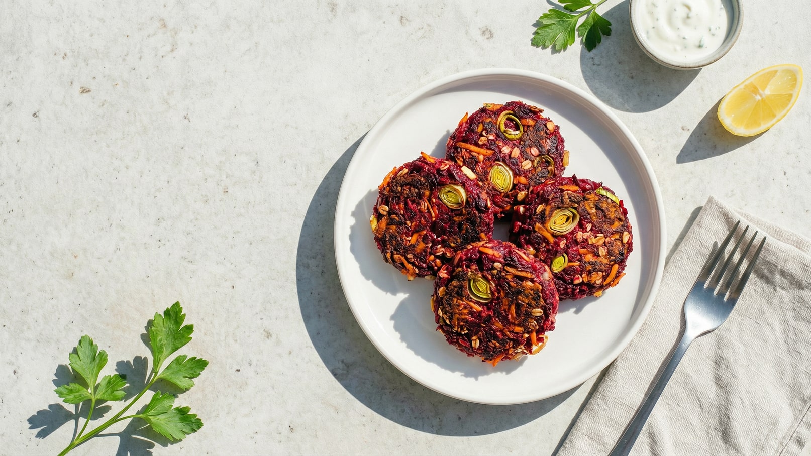 Auf einem weißen Teller liegen vier bunte Gemüsepattys aus Roter Beete und Karotten, garniert mit Lauchringen. Daneben sind ein kleiner Schälchen mit Joghurt und ein Stück Zitrone zu sehen. Ein Besteck liegt bereit, um das Gericht zu genießen.