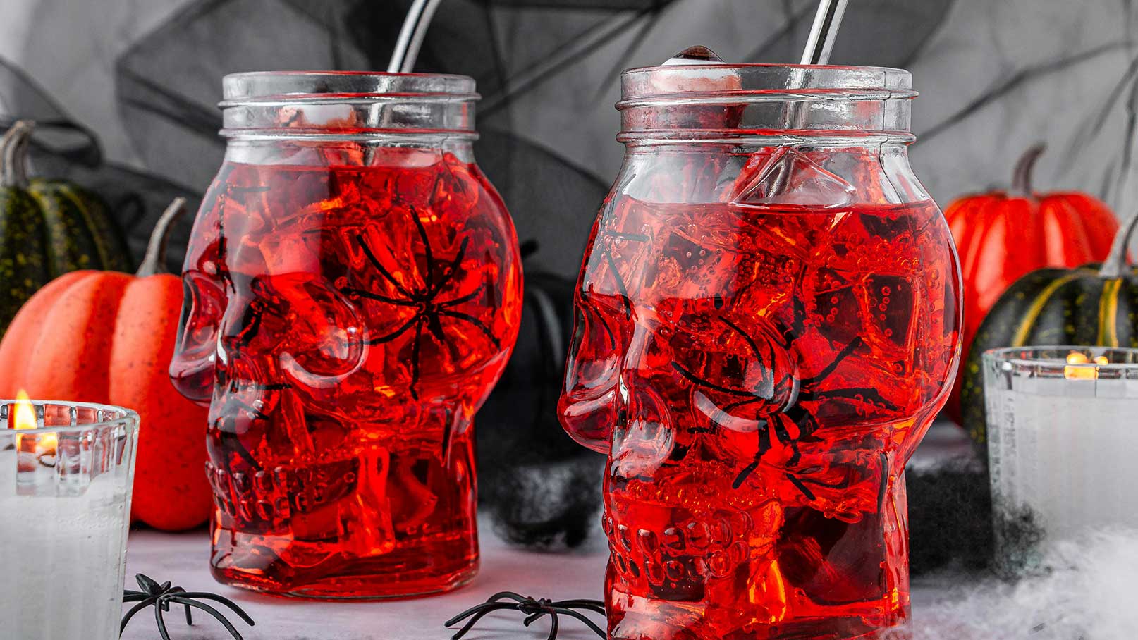 Zwei große Gläser in Schädel-Form enthalten ein rotes Getränk mit Eiswürfeln und schwarzen Spinnenmotiven. Im Hintergrund sind orangefarbene und grüne Kürbisse sowie Kerzen zu sehen. Die Atmosphäre ist festlich und erinnert an Halloween.