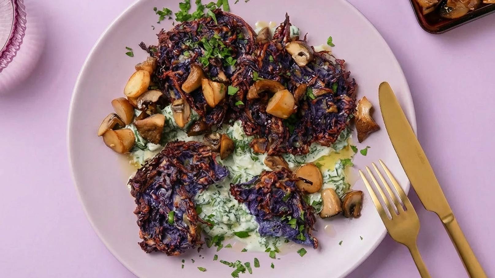 Ein Teller mit lila Frittiertem, umgeben von gebratenen Champignons und einer grünen, cremigen Beilage. Frische Kräuter sind darüber gestreut. Das Geschirr ist elegant, mit einem goldenen Besteck und einem pastellfarbenen Untergrund.