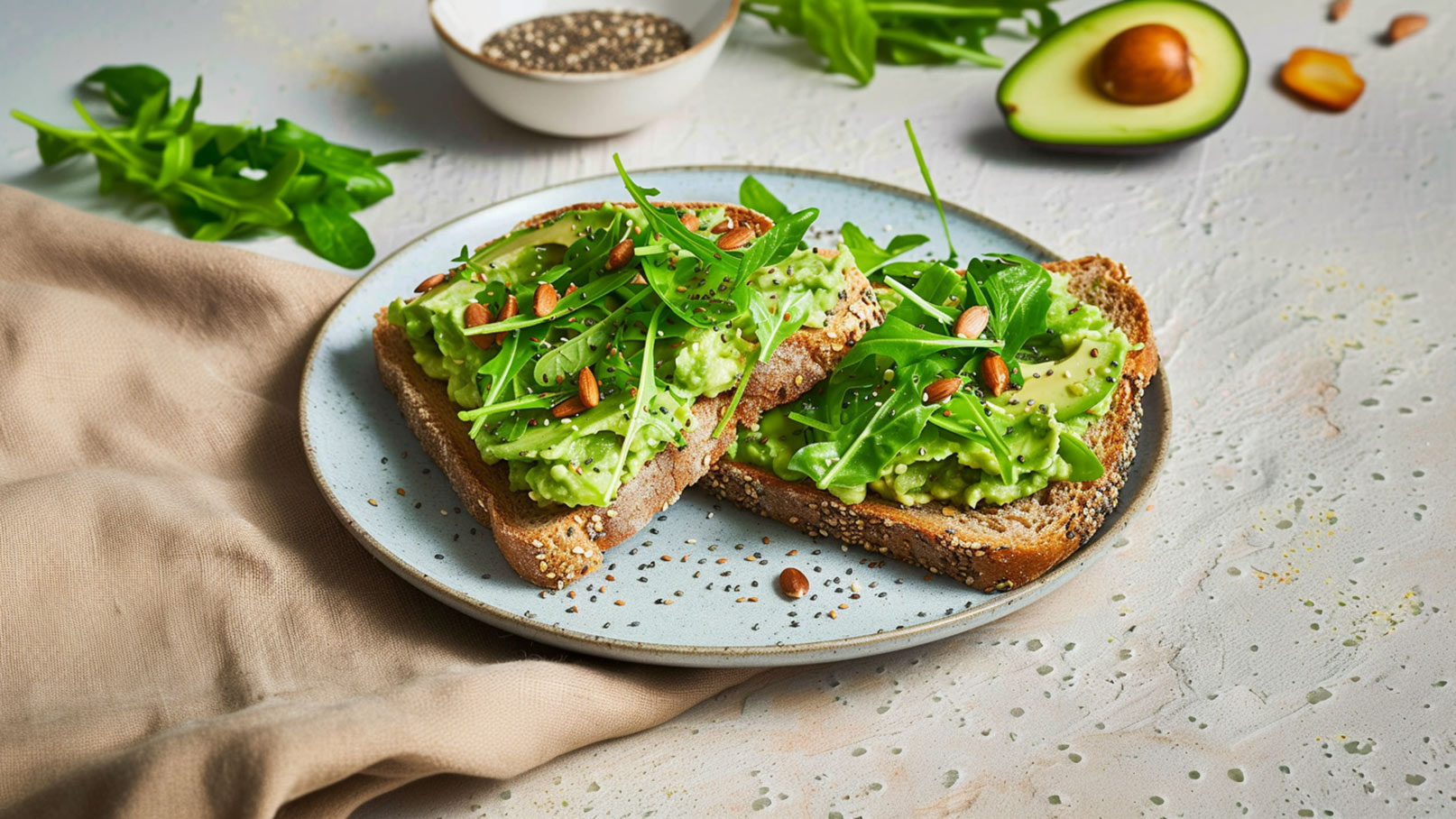 Zwei Scheiben Vollkornbrot auf einem blauen Teller, belegt mit einer cremigen Avocado-Mischung und frischem Rucola. Darüber sind einige geröstete Mandeln und Gewürze verteilt. Im Hintergrund ist eine halbe Avocado und ein kleiner Schälchen mit Chiasamen zu sehen.