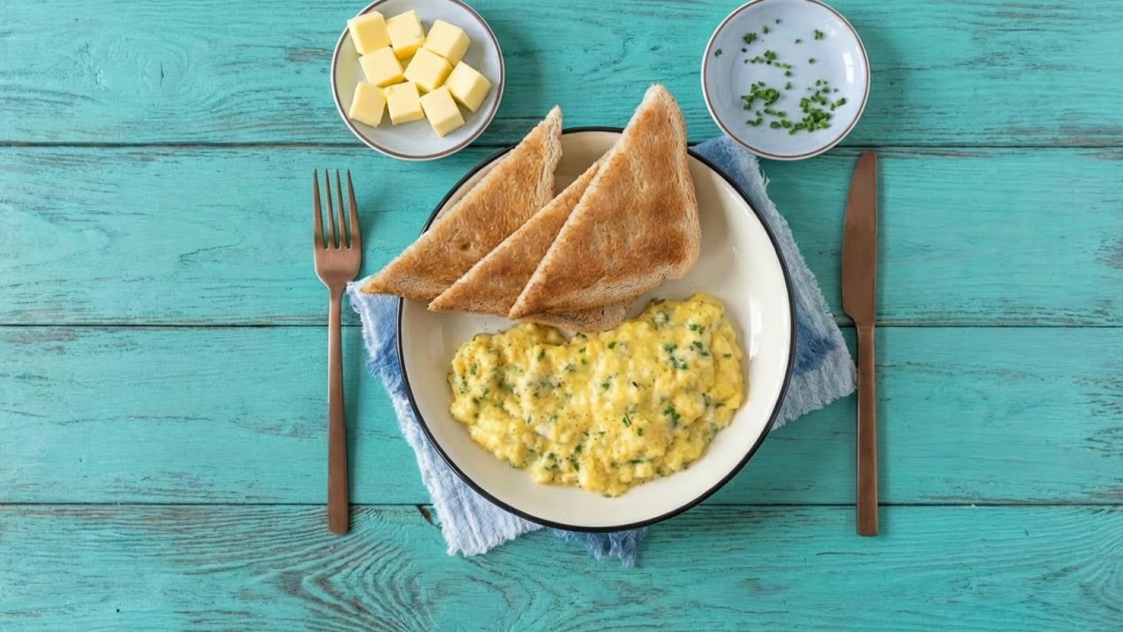 Ein Teller mit Rührei, das mit Kräutern garniert ist, liegt in der Mitte. Daneben befinden sich zwei Scheiben geröstetes Brot. Auf einem kleinen Teller sind Butterwürfel und ein weiterer Teller mit frischen Kräutern. Der Hintergrund ist in einem hellen Türkis gehalten.