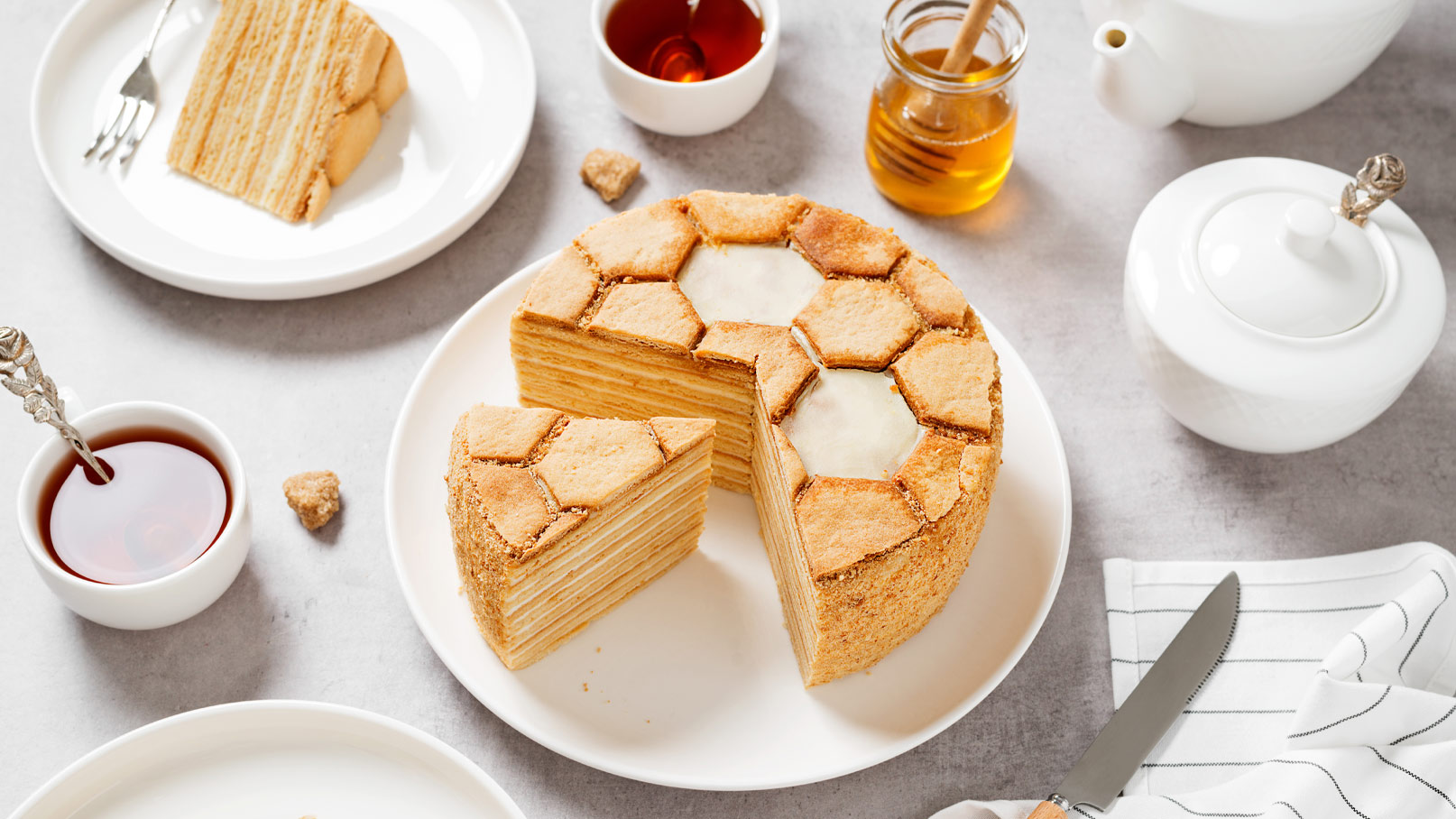 Ein runder, geschichteter Kuchen mit einer honigähnlichen Oberfläche, die in sechseckige Formen geschnitten ist. Ein Stück des Kuchens wurde herausgenommen, und der Kuchen liegt auf einem weißen Teller. Daneben befinden sich eine Tasse Tee, ein Honigglas und mehrere Geschirrteile.