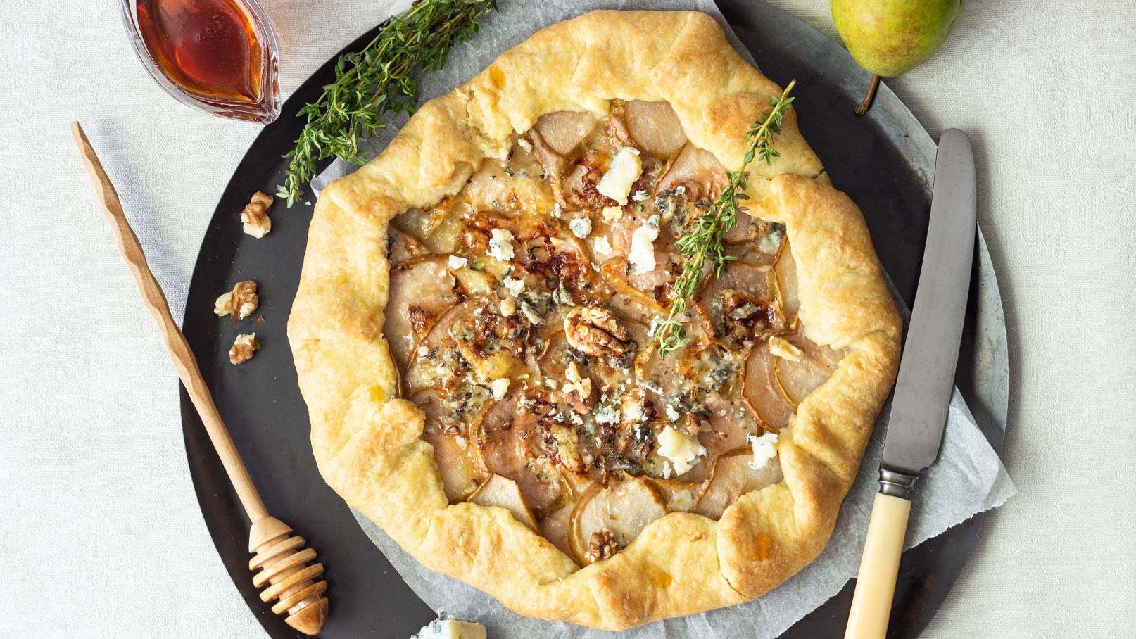 Ein frisch gebackener, rustikaler Kuchen mit einer goldbraunen Kruste, gefüllt mit dünn geschnittenen Birnen, Walnüssen und Käse. Oben sind einige Thymianzweige verteilt. Daneben befindet sich ein Honiglöffel und eine kleine Schale mit Sirup.