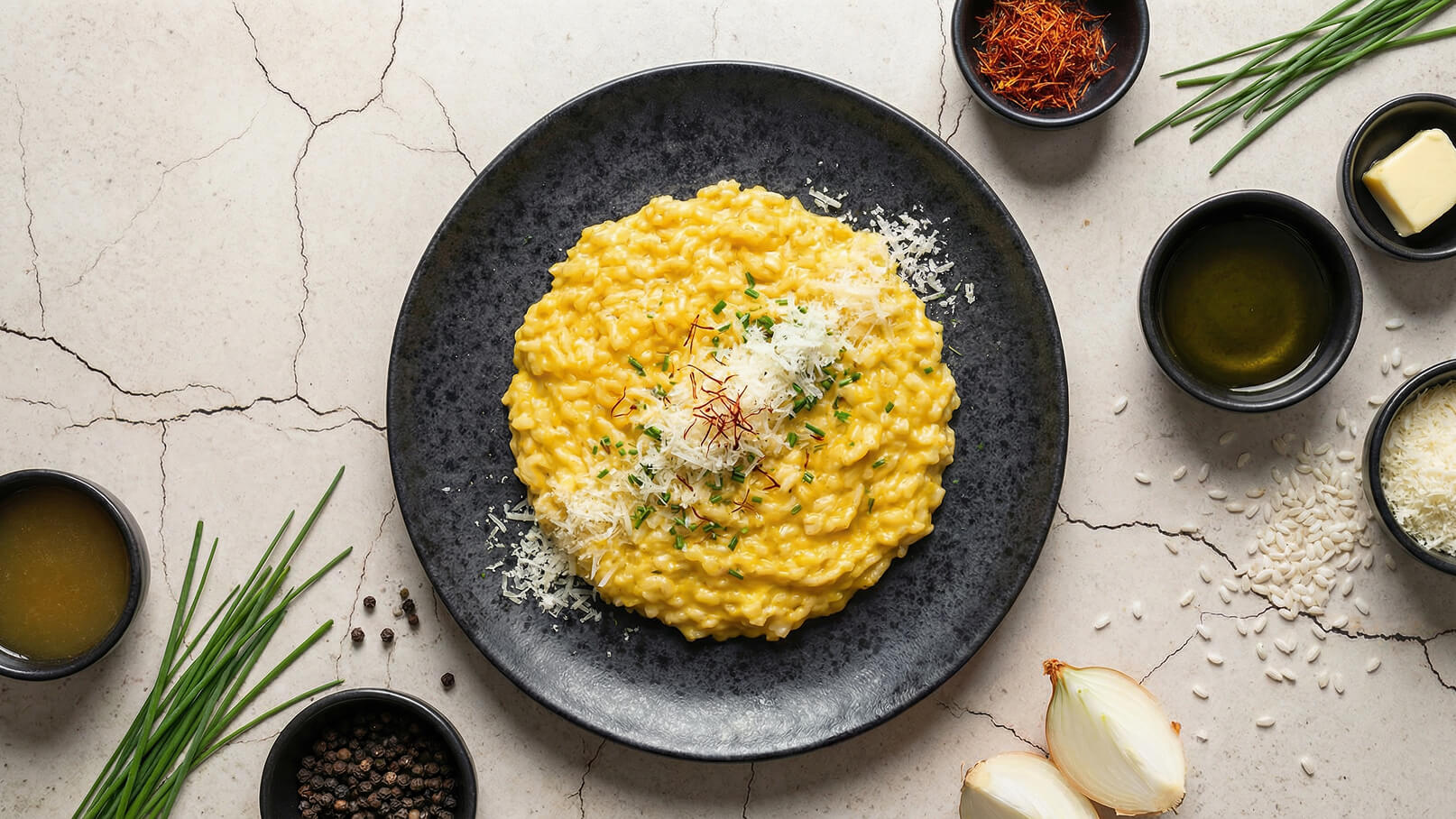 Ein schwarzer Teller mit cremigem, gelbem Risotto, garniert mit Frischkäse und einigen roten Safranfäden. Um den Teller herum sind verschiedene Zutaten wie Schnittlauch, Gemüsebrühe und Gewürze angelegt. Der Hintergrund besteht aus einer hellen, marmorierten Oberfläche.