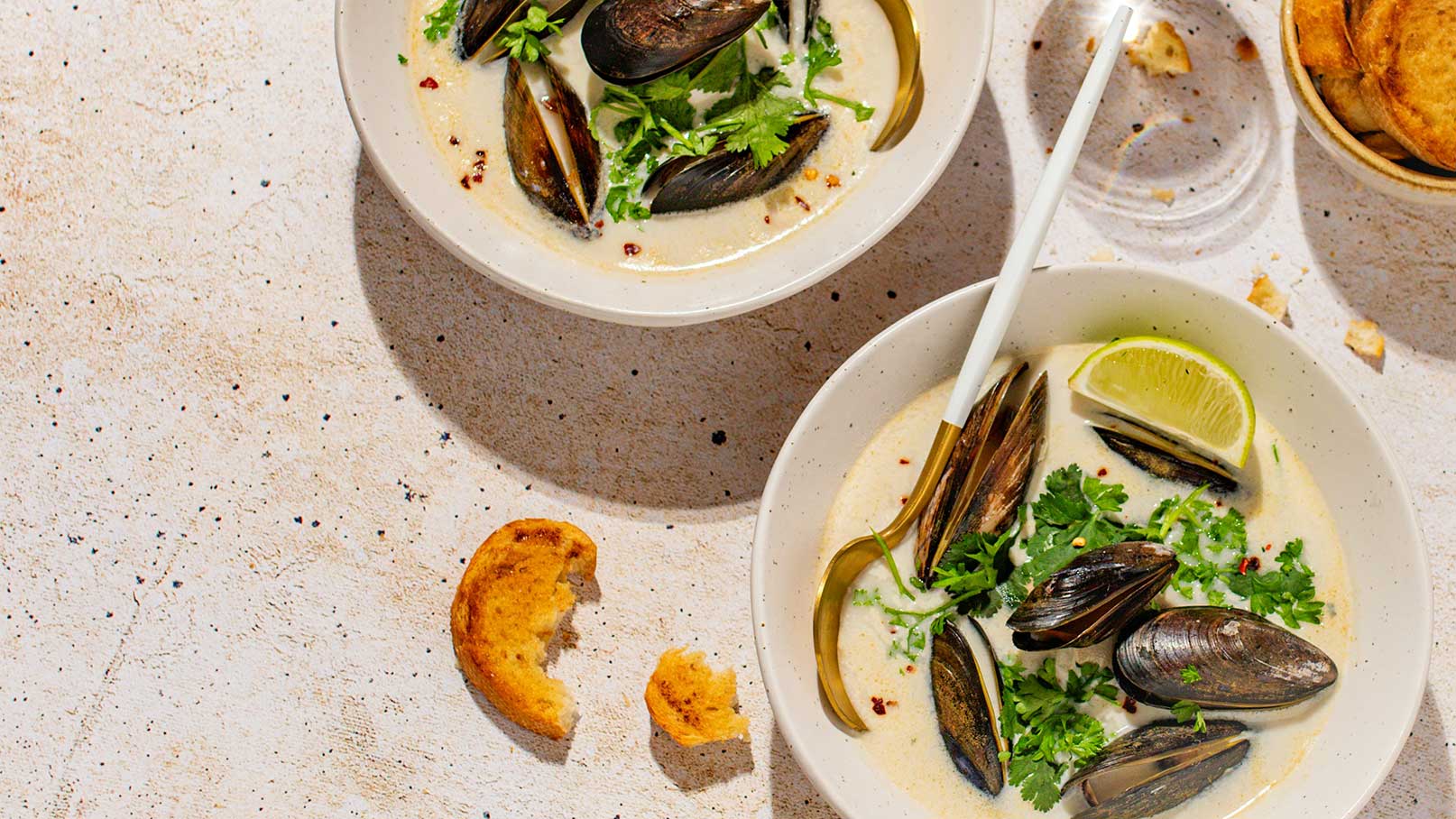 Zwei Schalen mit cremiger Muschelsuppe, garniert mit frischem Koriander und Limettenscheiben. Neben den Schalen stehen Gabeln und ein Glas Weißwein. Im Hintergrund sind einige knusprige Chips zu sehen.