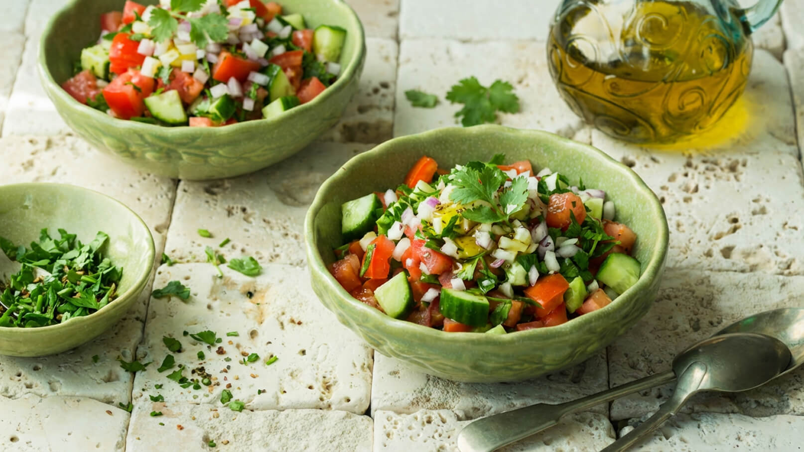 Zwei Schalen mit frischem Salat stehen auf einem Tisch. Der Salat besteht aus gewürfelten Tomaten, Gurken, Zwiebeln und frischen Kräutern. Neben den Schalen liegt eine kleine Menge gehackte Kräuter auf dem Tisch, und es ist eine Flasche Öl in der Nähe zu sehen.