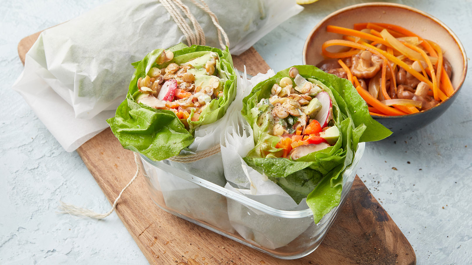 Zwei frische, grüne Salatwraps gefüllt mit buntem Gemüse und Saaten stehen in einem durchsichtigen Behälter. Daneben befindet sich eine Schale mit eingelegtem Gemüse. Die Szene vermittelt frische und gesunde Ernährung.